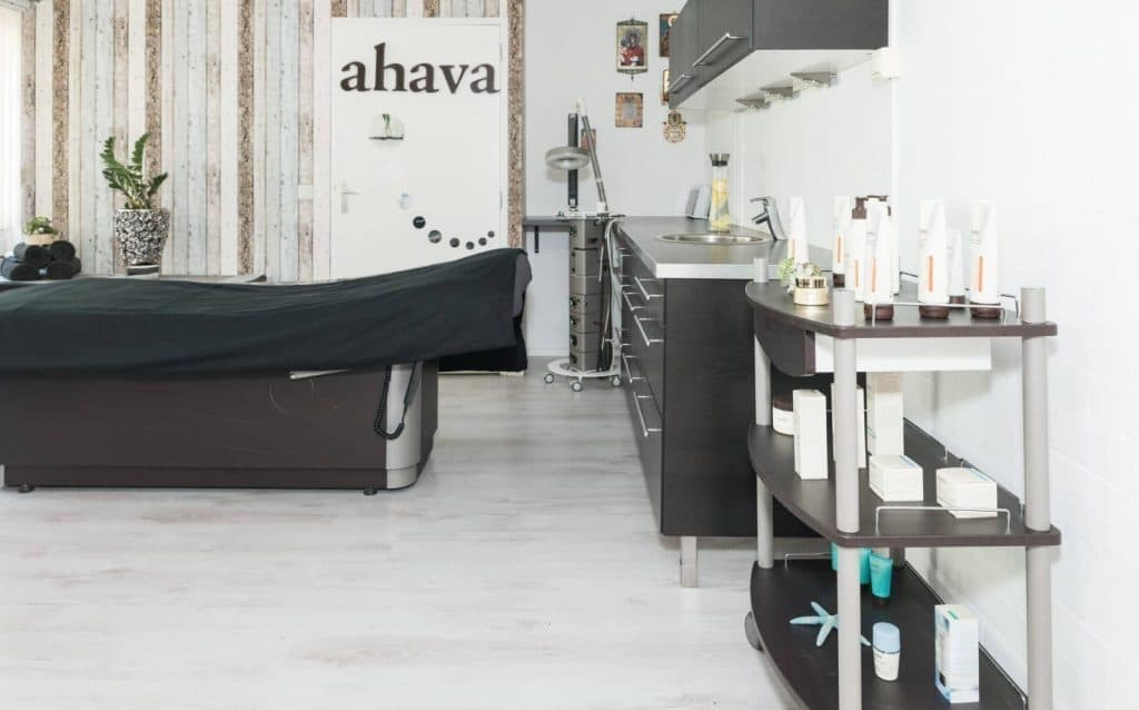De lichte behandelkamer met behandelbed en verzorgingsproducten; op de deur staat AHAVA